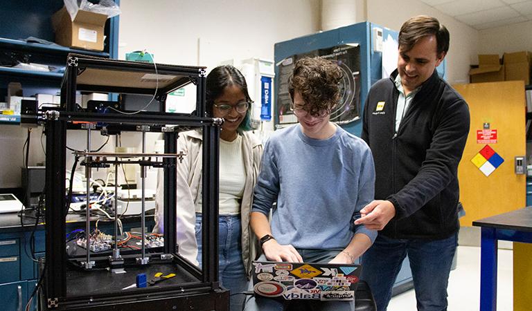 Steven Santana and two students work together on a laptop near a printing machine.